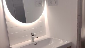 A bathroom with a white sink, a mirror with a circular light, and a wooden cabinet. - Authentique Bouquet (Le Mans)