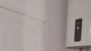 A laundry room with a front-loading washing machine, a shelf with a green container, and a wall-mounted device above it. - Authentique Bouquet (Le Mans)
