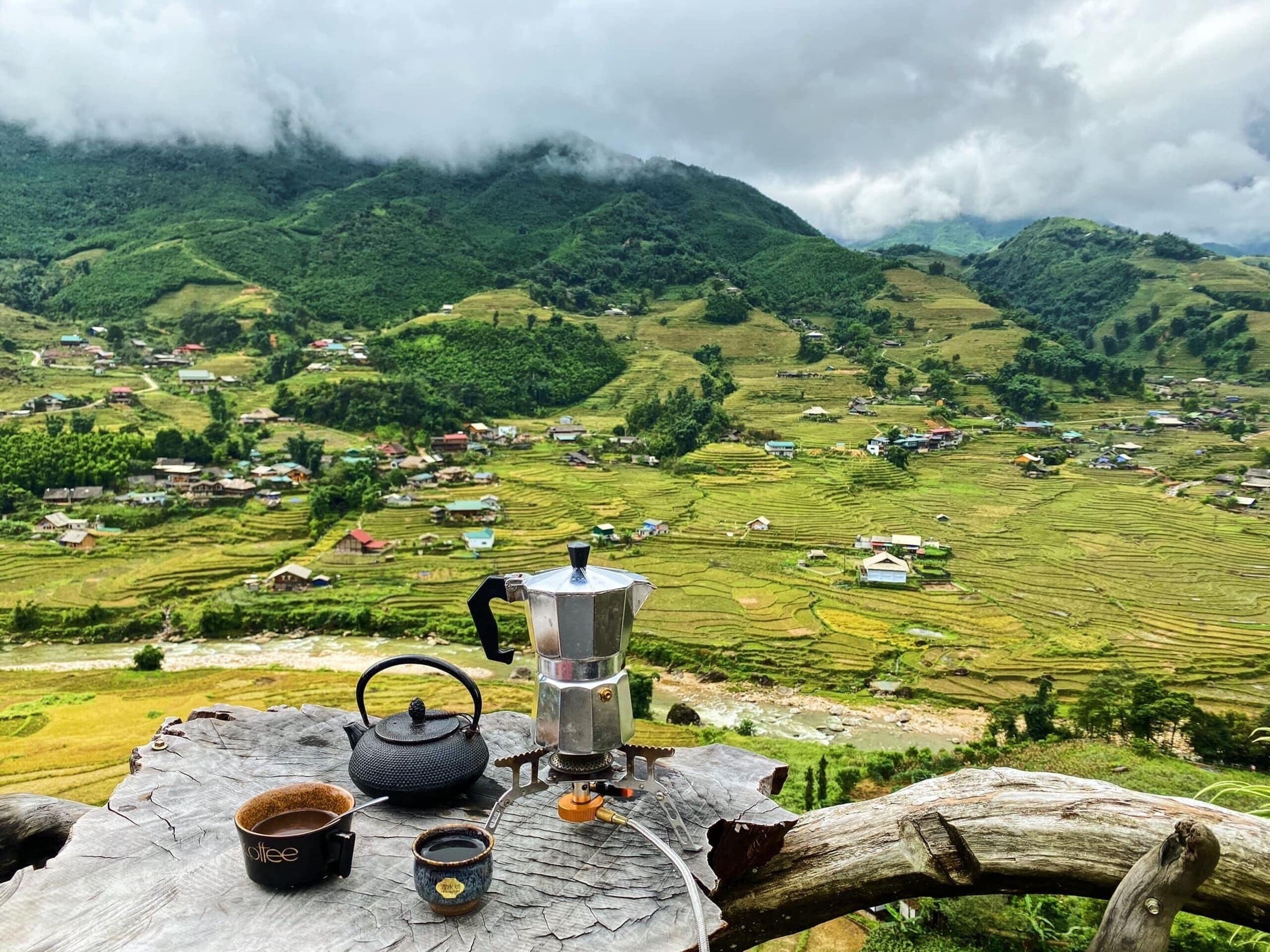 Entorno rústico al aire libre con una mesa que presenta un hervidor, tazas y una estufa portátil, con vistas a campos escalonados y un pueblo.