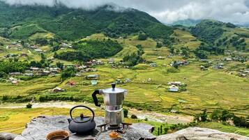 Entorno rústico al aire libre con una mesa que presenta un hervidor, tazas y una estufa portátil, con vistas a campos escalonados y un pueblo.