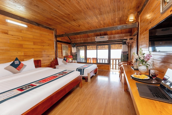 A wooden cabin room with two beds, a dining table with a laptop, and a view of the outdoors.