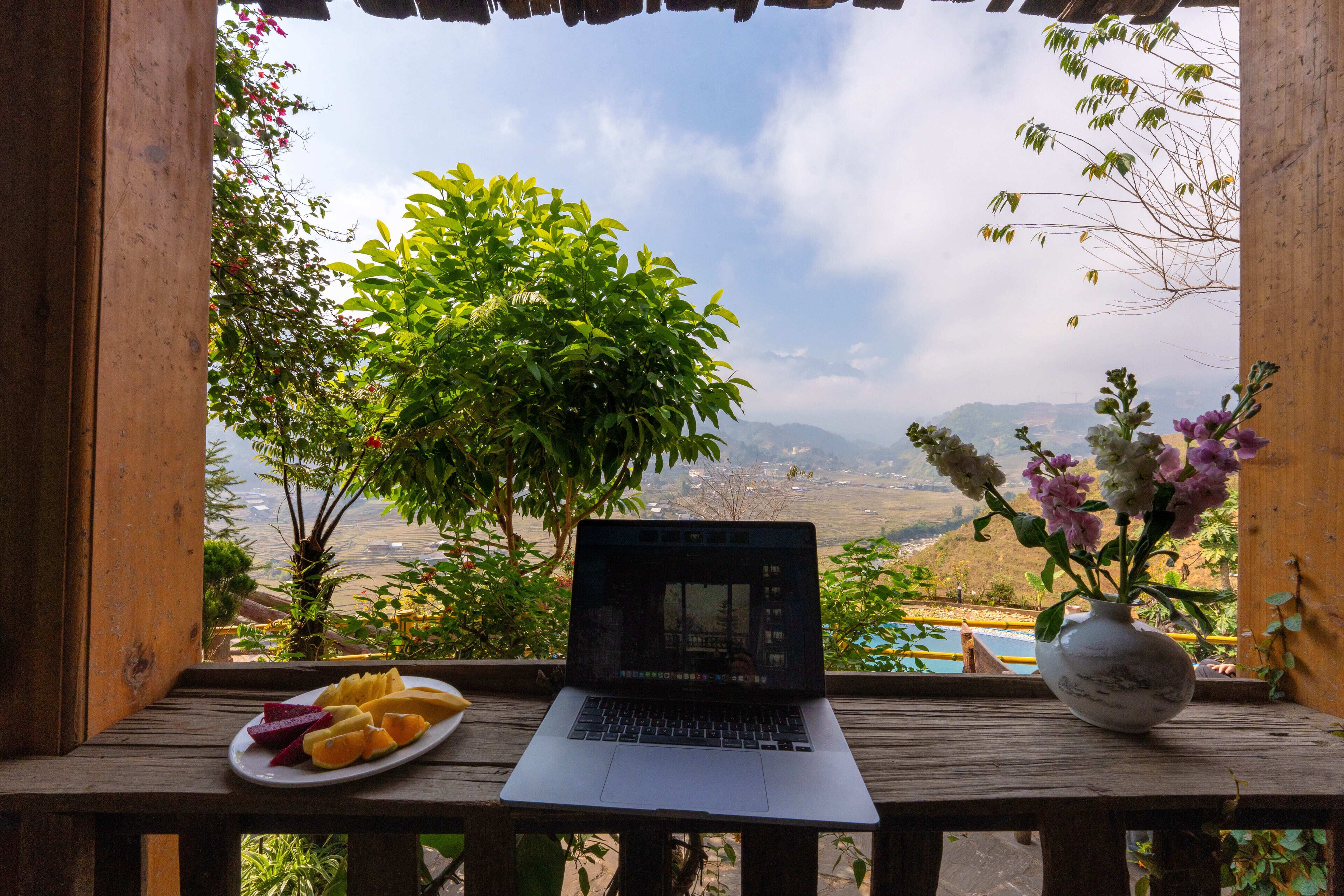 Una computadora portátil sobre una mesa de madera con un plato de fruta y un florero, con vistas a un paisaje pintoresco.