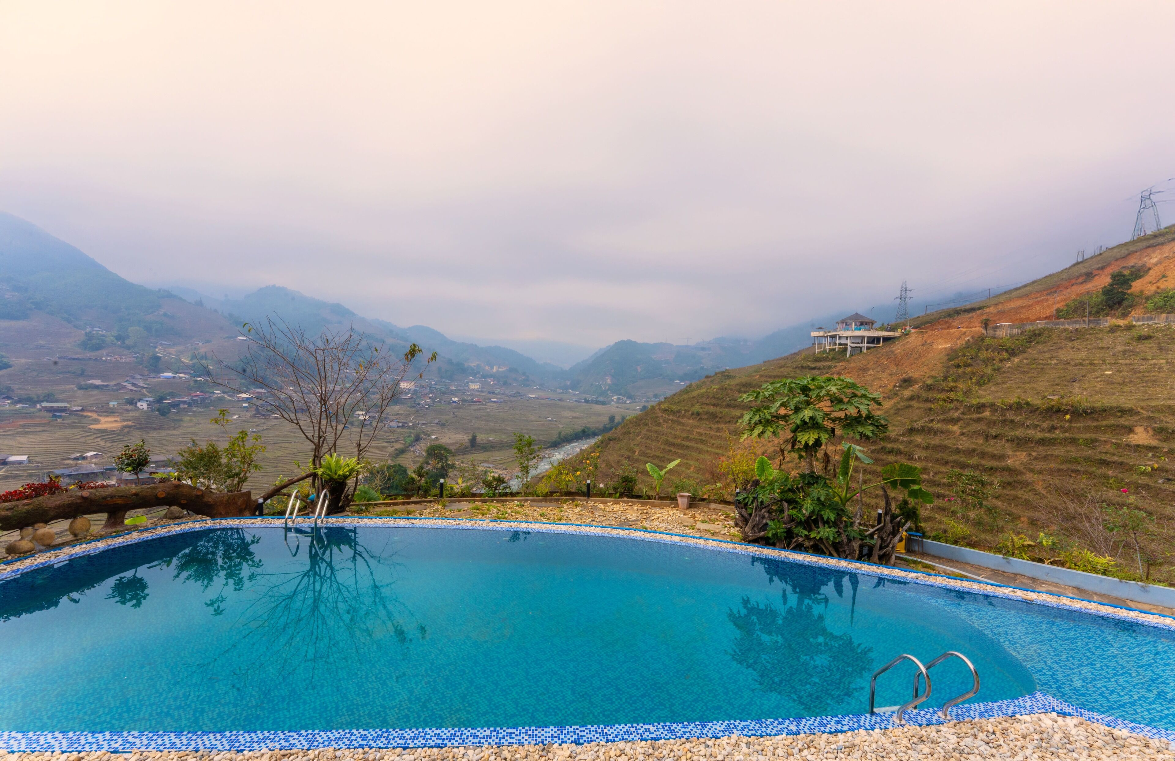 A pool with clear blue water, surrounded by pebbles and plants, overlooking a valley with distant hills and a building in the background.