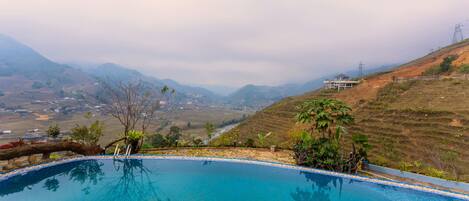 A pool with clear blue water, surrounded by pebbles and plants, overlooking a valley with distant hills and a building in the background.