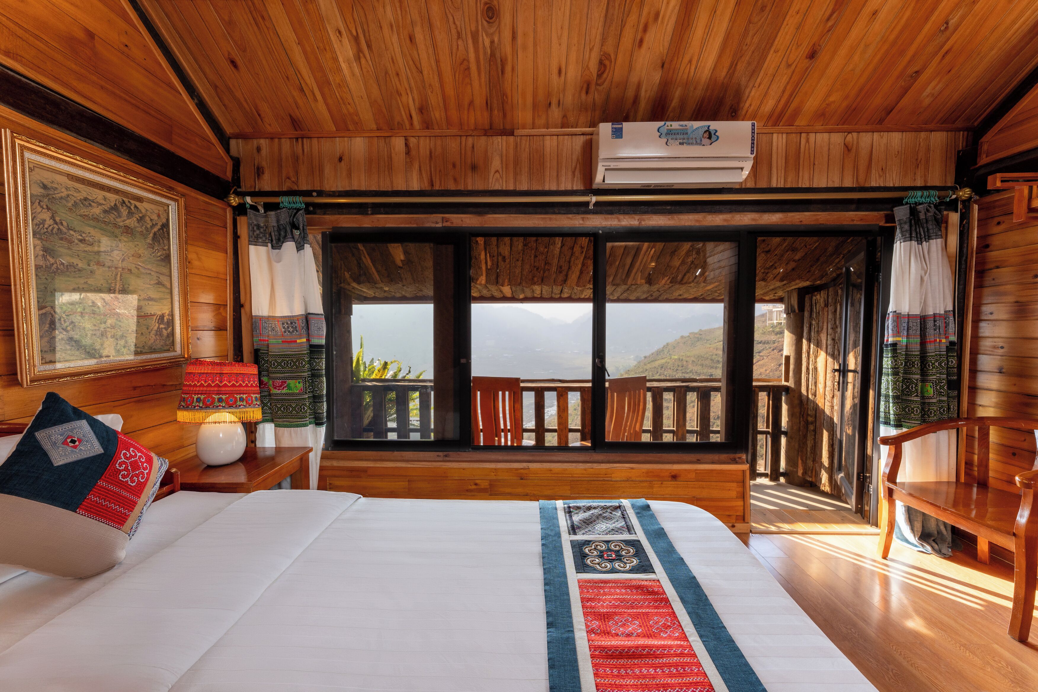 Habitación de cabaña de madera con una cama, una mesita de noche, una lámpara y un ventanal con vistas a las montañas.