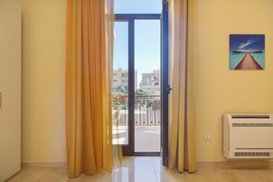 Una habitación con puerta de vidrio corredera que da a un balcón, aire acondicionado instalado en la pared y un cuadro con una playa.