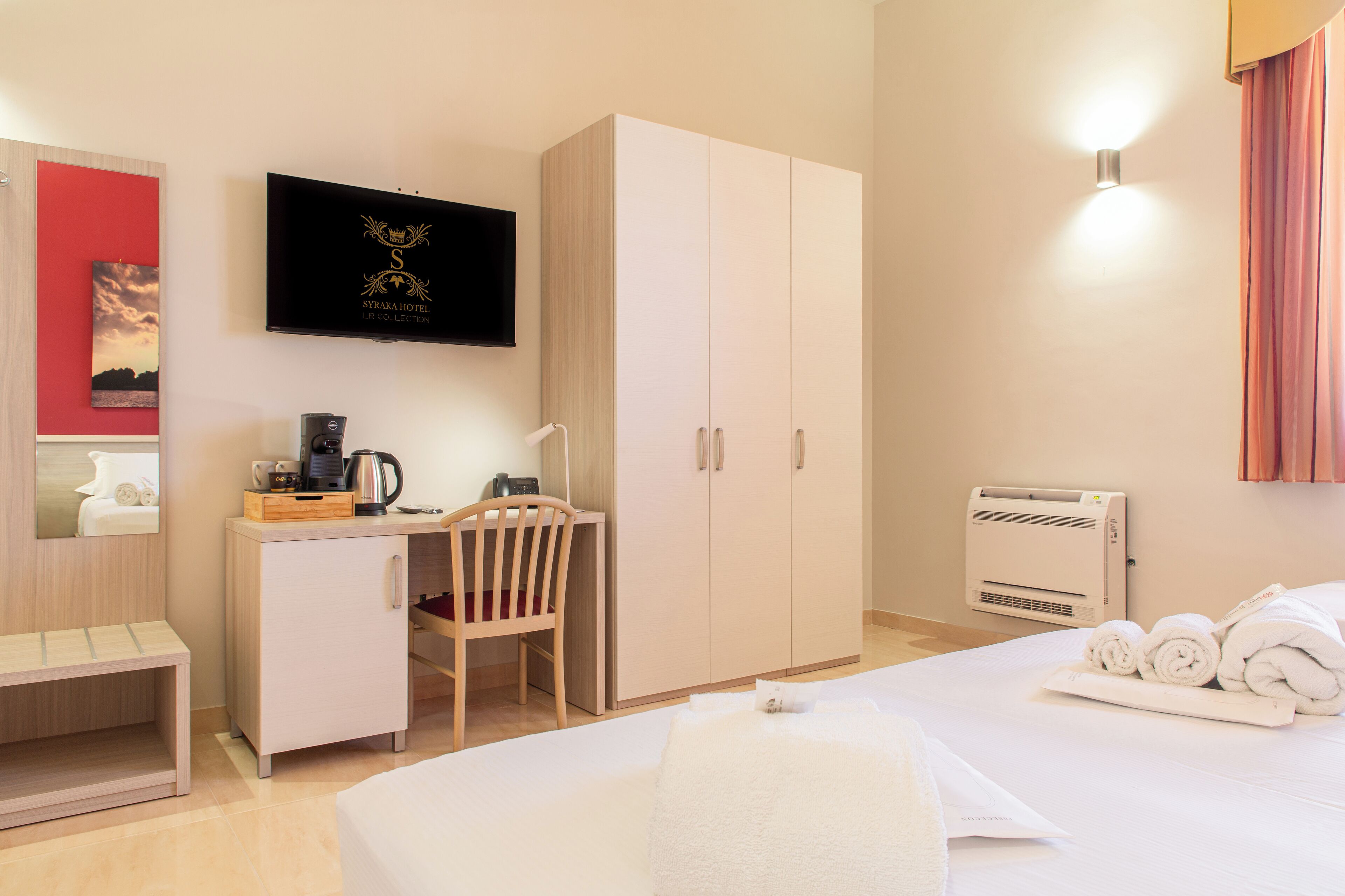A hotel room with a bed, a desk with a chair, a wardrobe, a television, and a wall-mounted heater.