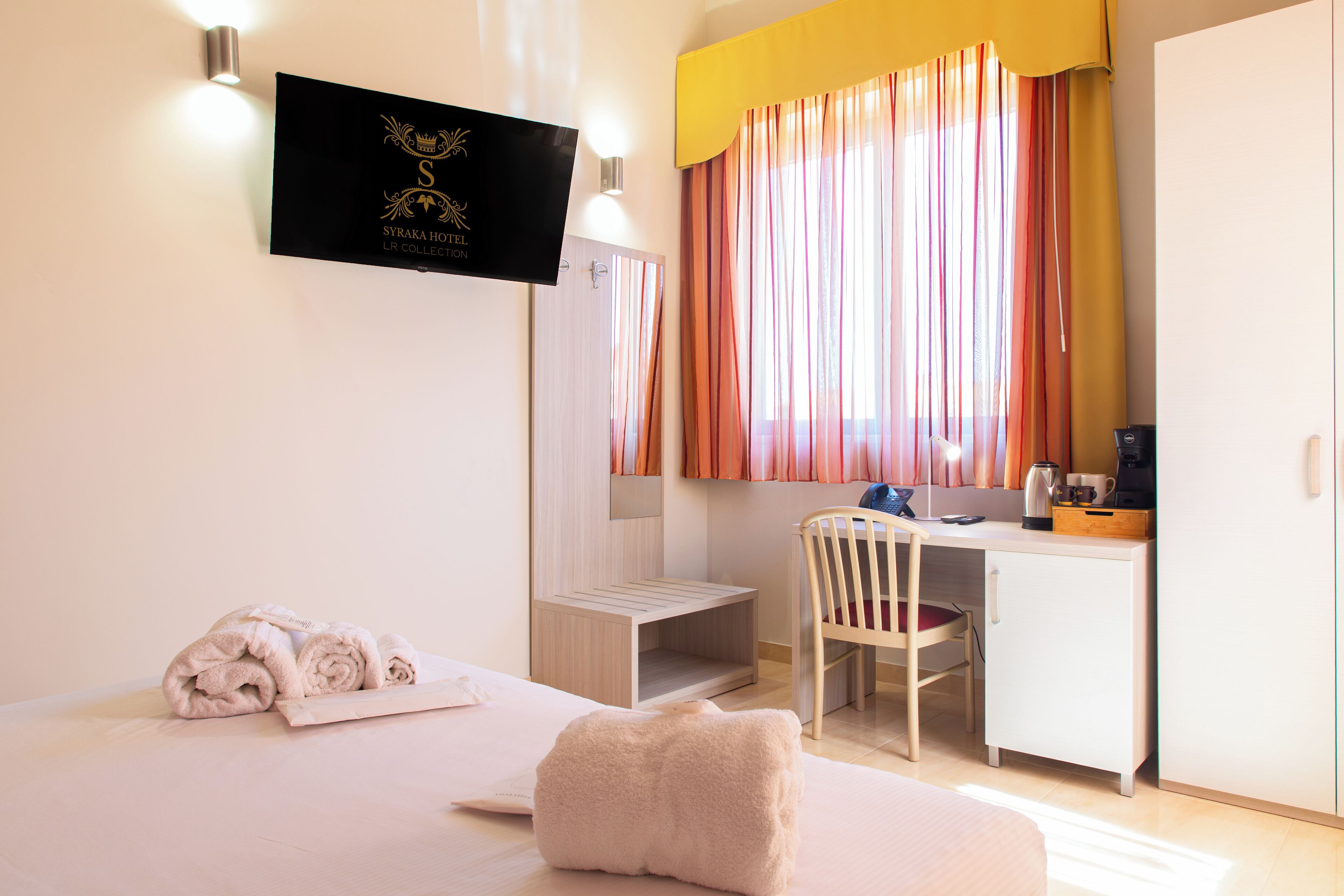 A hotel room with a bed, a desk, a chair, a small table, a coffee maker, and a TV on the wall.