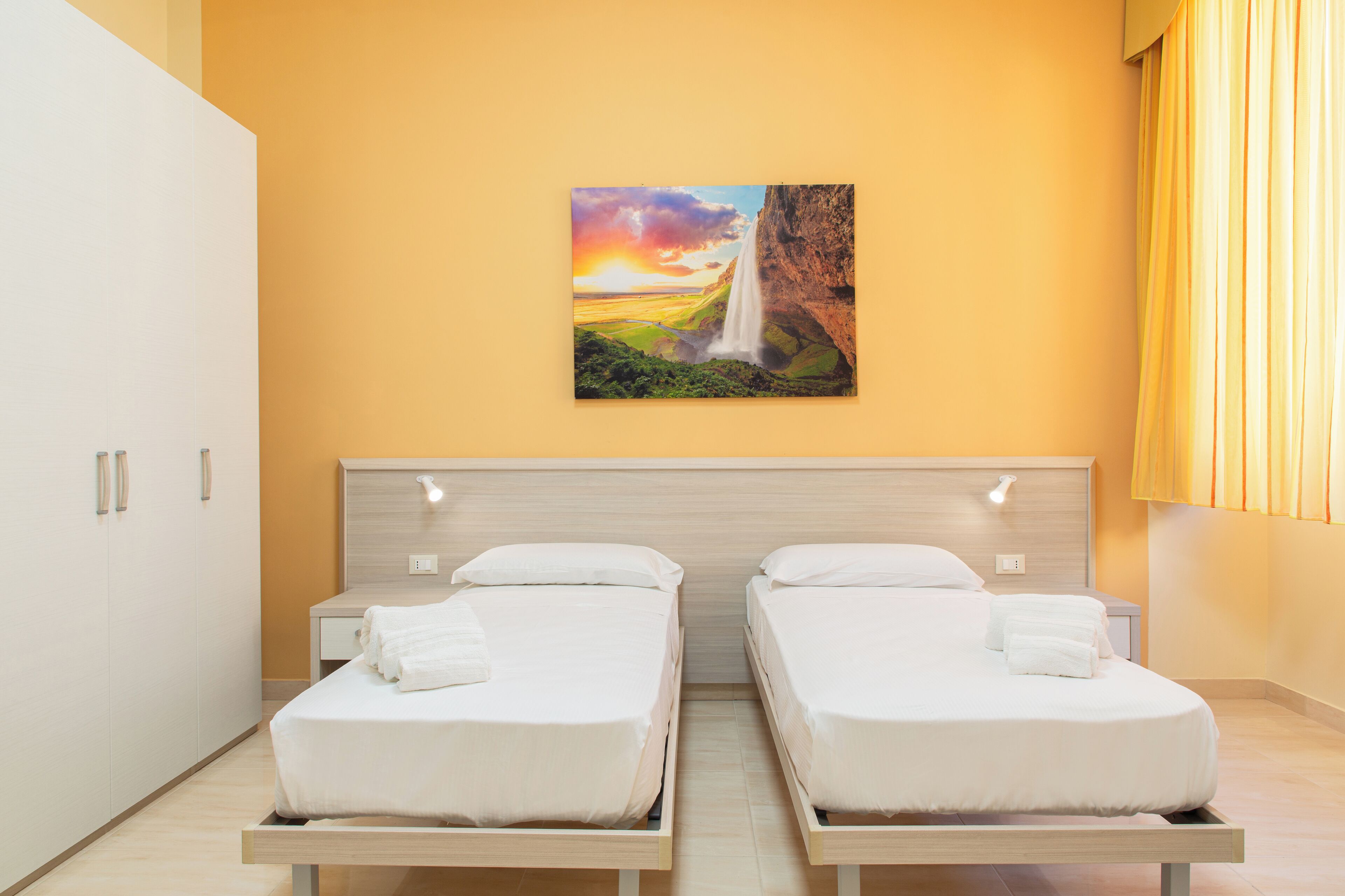 A hotel room with two single beds, a wooden headboard, a painting of a waterfall, and a yellow wall.
