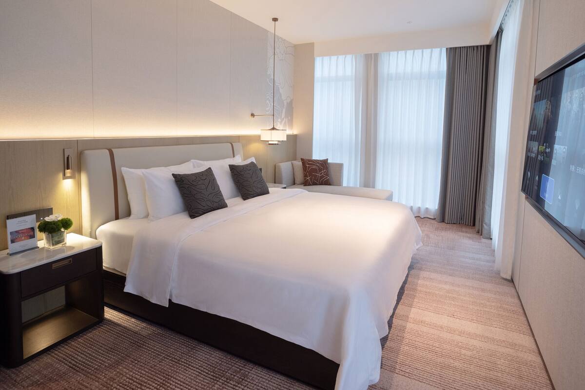 Habitación de hotel con una cama grande, una mesita de noche con lámpara y vistas a la ciudad desde la ventana.