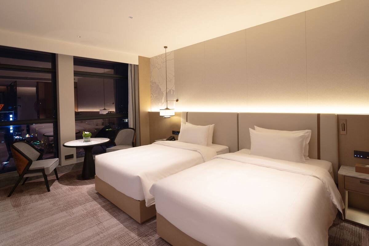 Habitación de hotel con dos camas, una mesita, una silla y vista a la ciudad de noche.