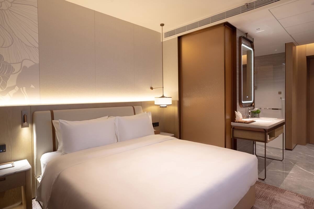Una habitación de hotel moderna con una cama grande, una mesita de noche, un escritorio con un espejo y una lámpara fijada a la pared.