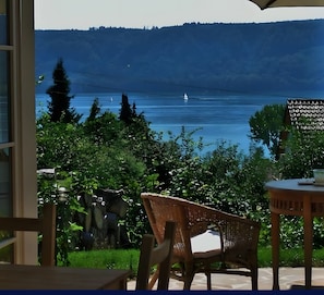Vista dalla finestra su un lago con una barca a vela, immerso nel verde e con una sedia.