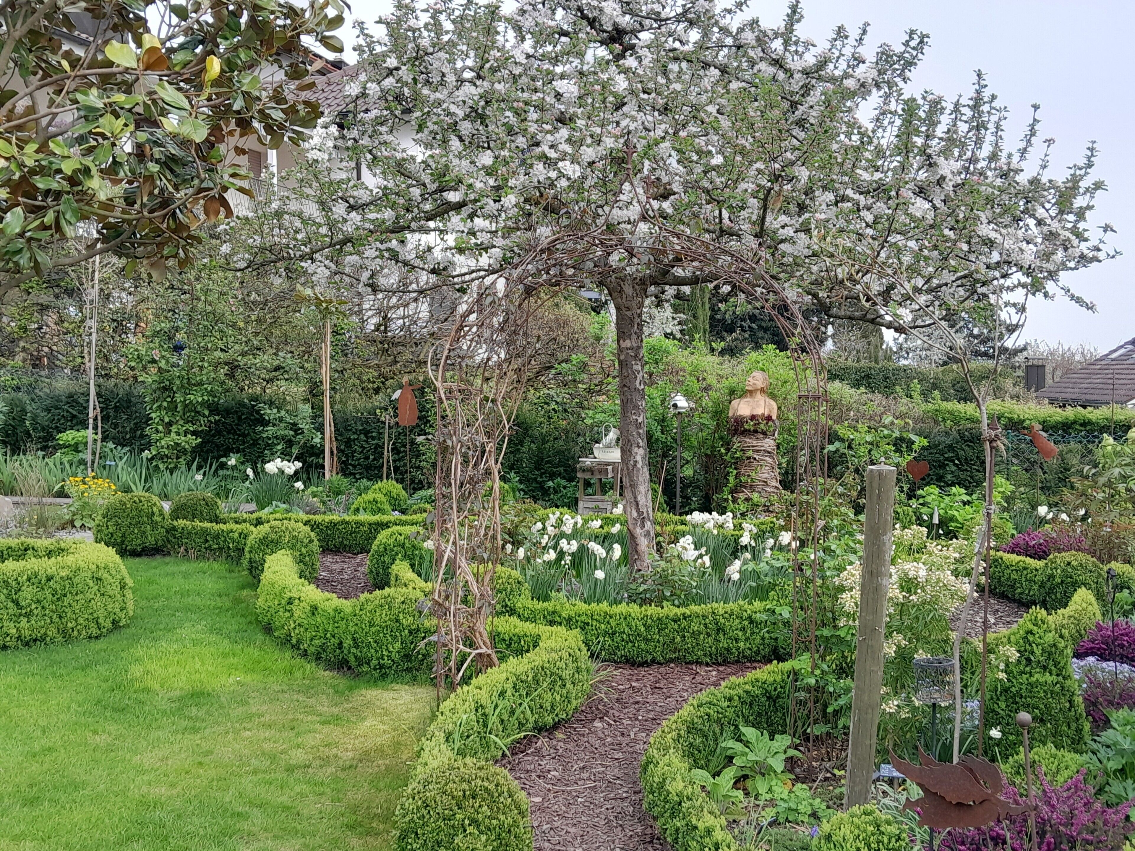 A garden with a variety of plants, a statue, and a pergola with flowers.