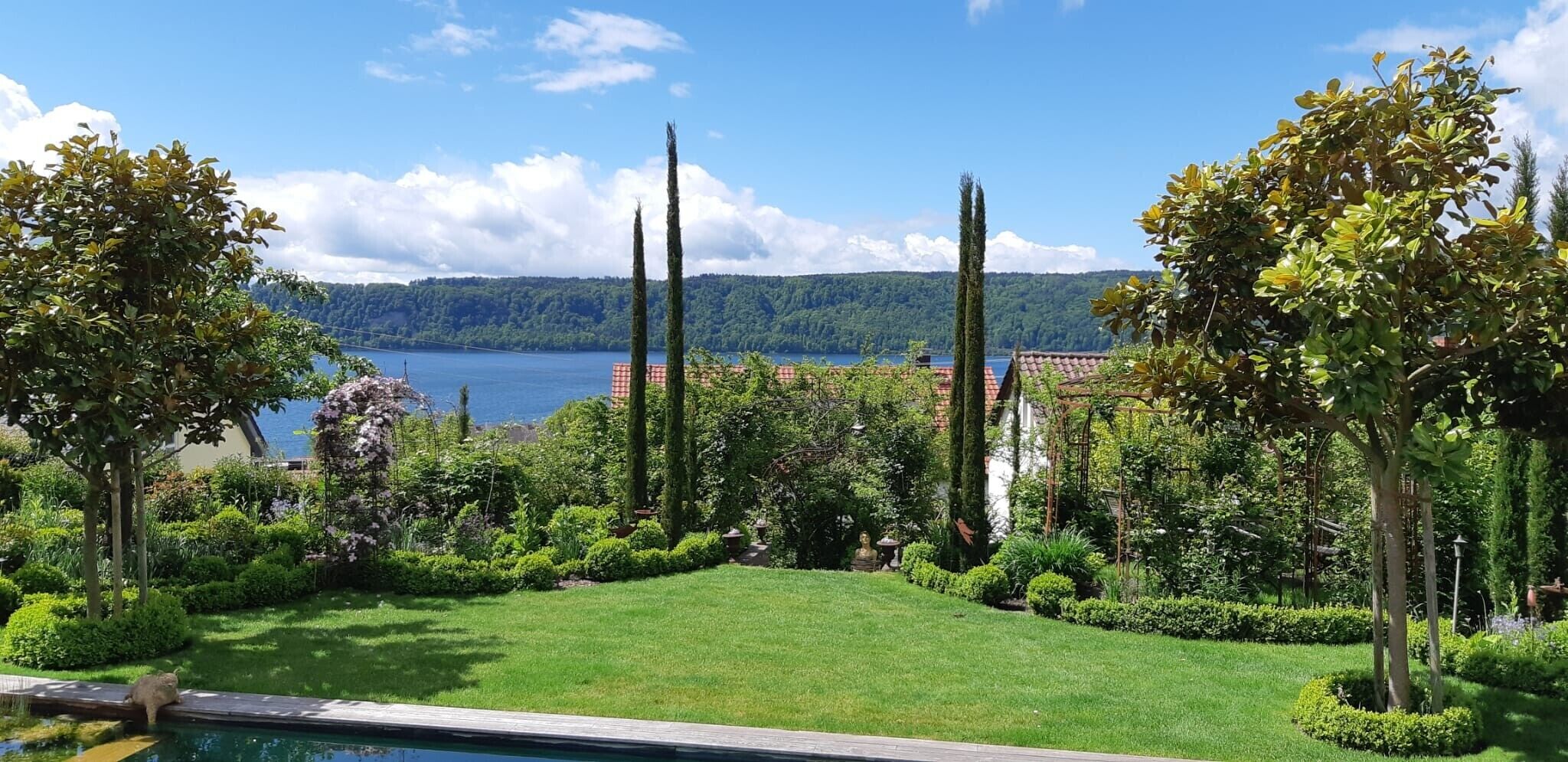 Un jardín bien cuidado con piscina, abundante vegetación y vistas a un lago y colinas distantes.