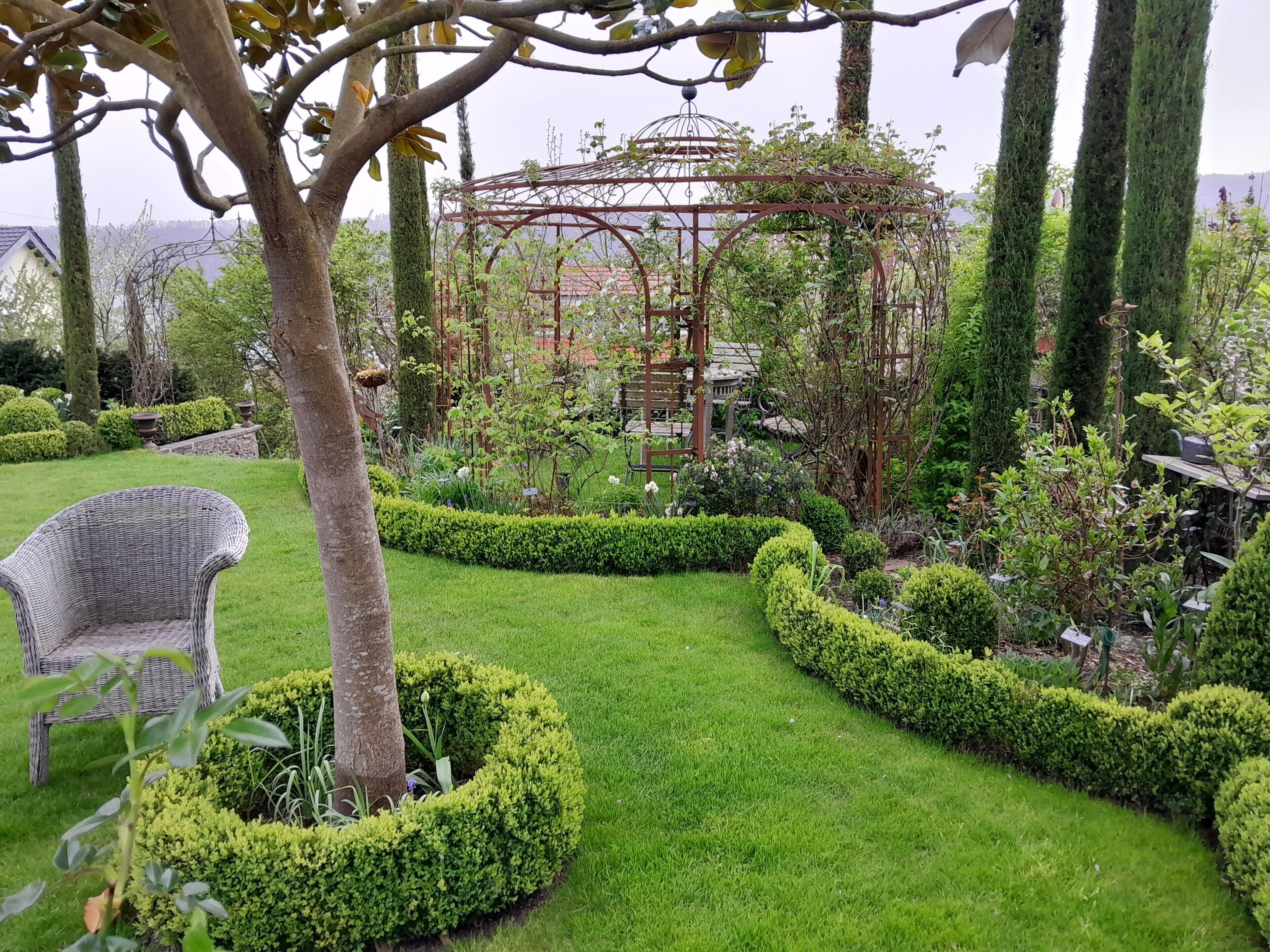 A garden with a pergola, neatly trimmed hedges, and a wicker chair.