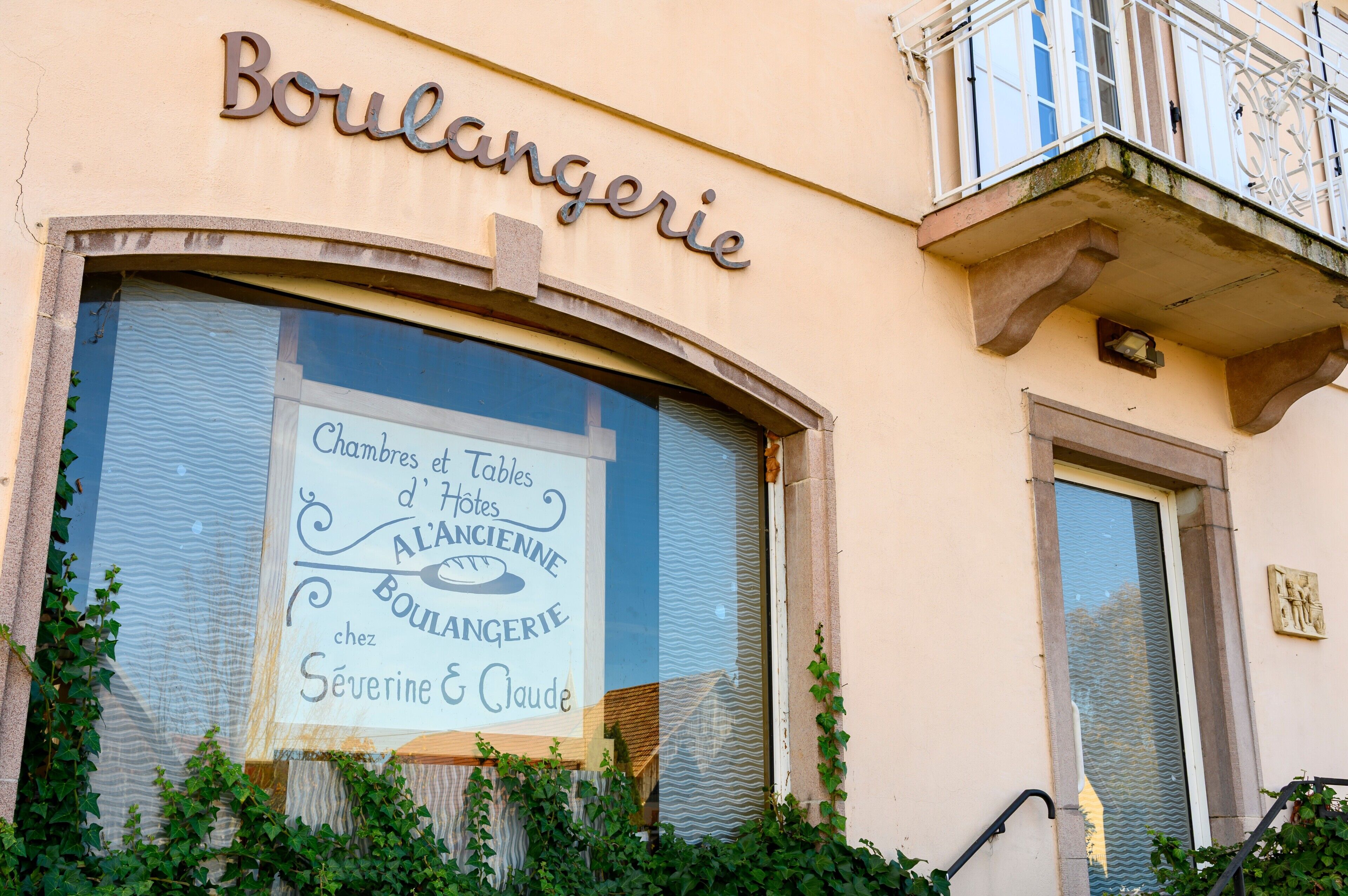 Un bâtiment avec une enseigne indiquant « Boulangerie » et une vitrine où est affiché un panneau publicitaire pour « Chambres et Tables d’hôtes à l’ancienne chez Boulangerie Séverine et Claude ».