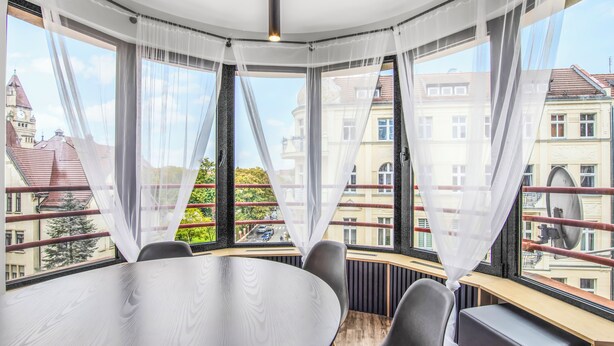 A corner room with a round table and chairs, large windows with sheer curtains, and a view of buildings and trees. - Apartamenty Sun & Snow Rozbrat Ołbin (Wrocław)