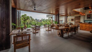 A spacious dining area with wooden furniture and large windows offering a view of greenery and buildings. - Lawangkala Resort Ubud (Ubud)
