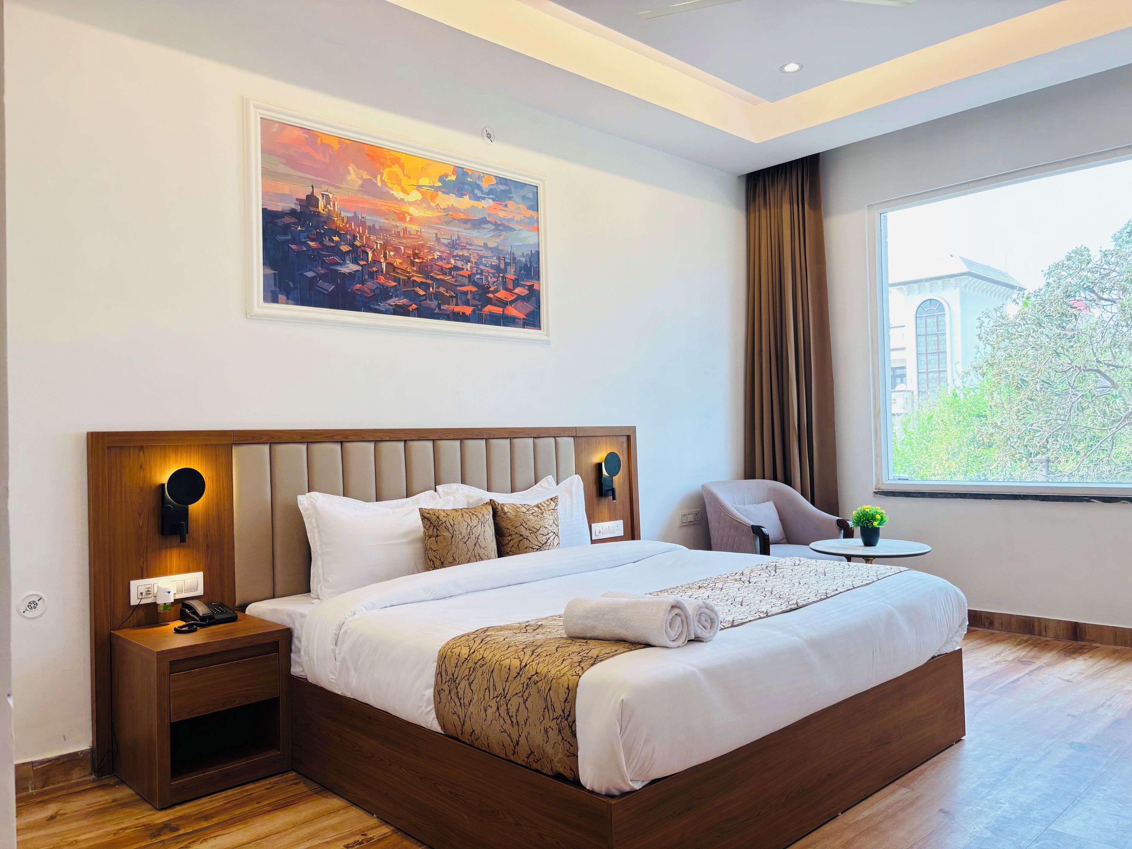 A hotel room with a large bed, a bedside table with a phone, a small table with a plant, a chair, and a painting on the wall.