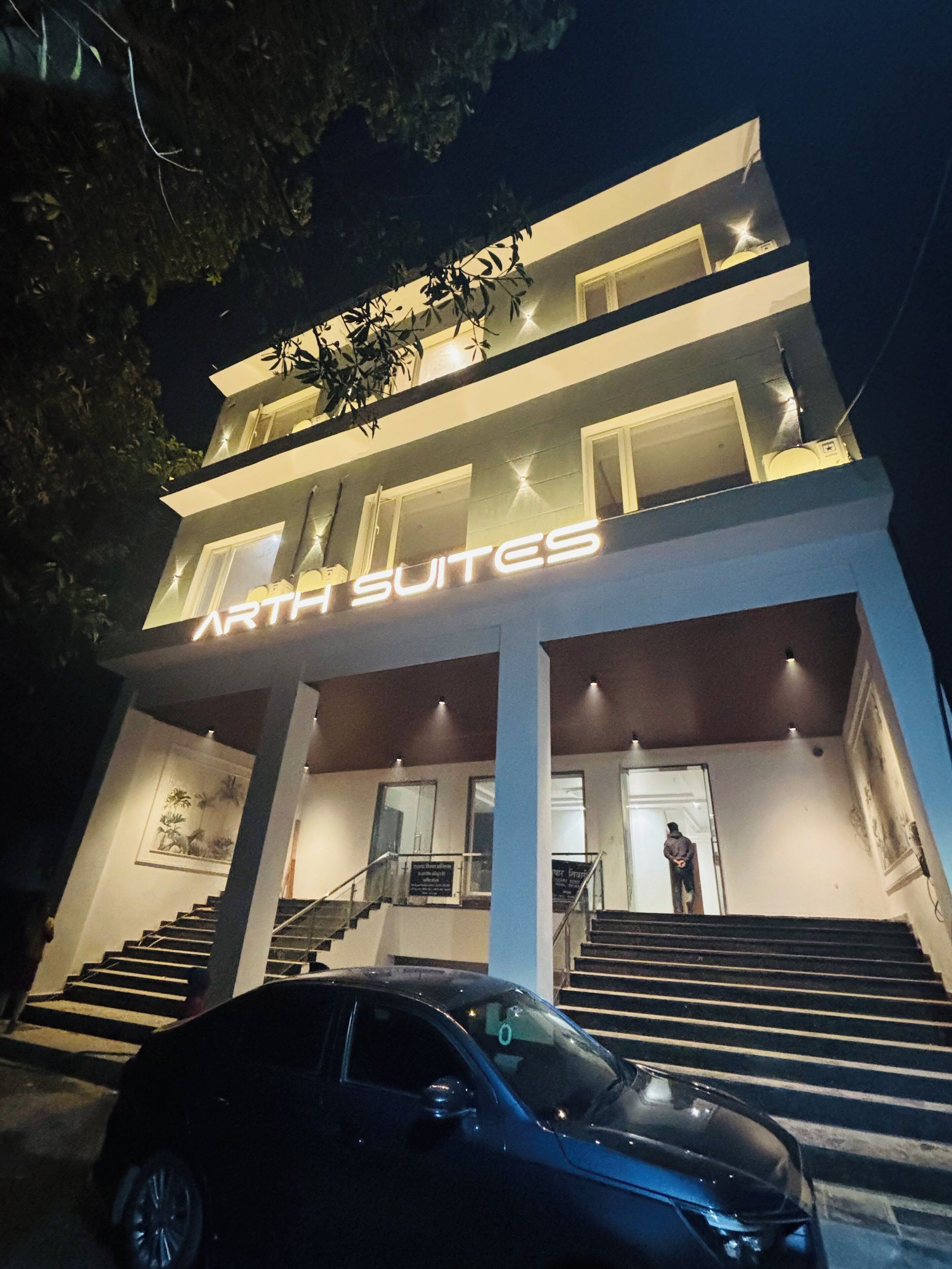 Un edificio moderno con un cartello che indica 'ARTH SUITES' all'ingresso, con una scala e un'auto parcheggiata di fronte.