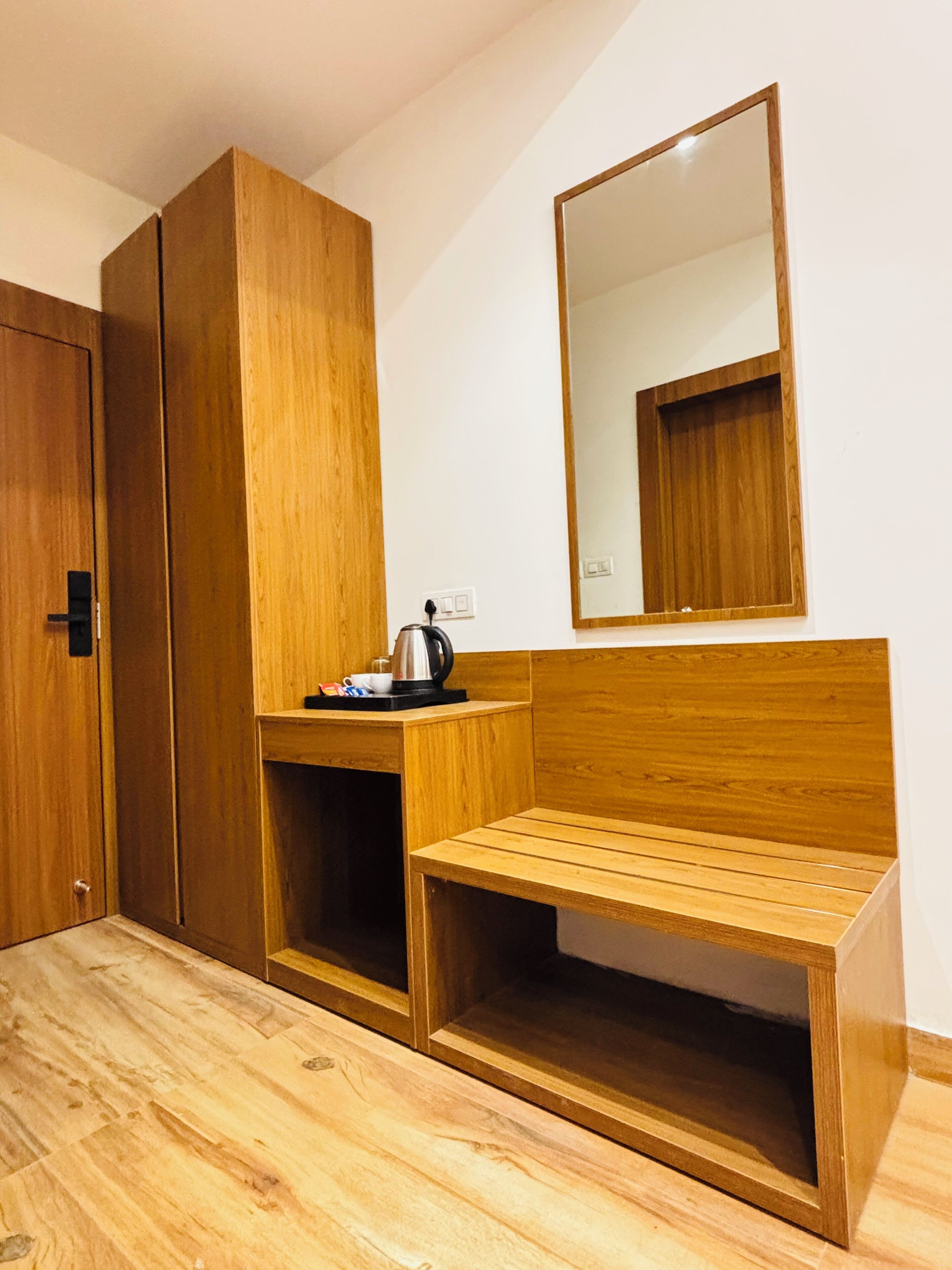 Una habitación con un armario de madera, una pequeña mesa de madera con un hervidor, un banco y un espejo en la pared.