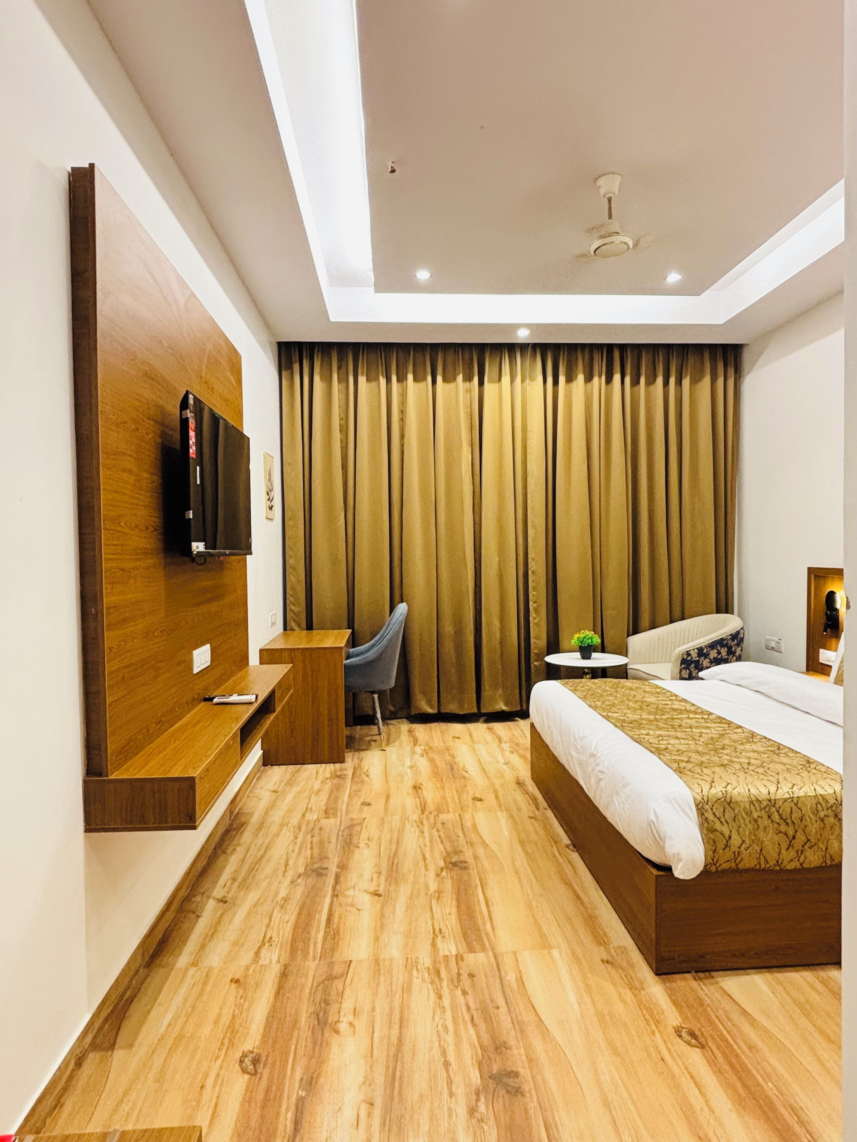 A hotel room with a wooden floor, a bed, a desk, a chair, a TV mounted on the wall, and a ceiling fan.