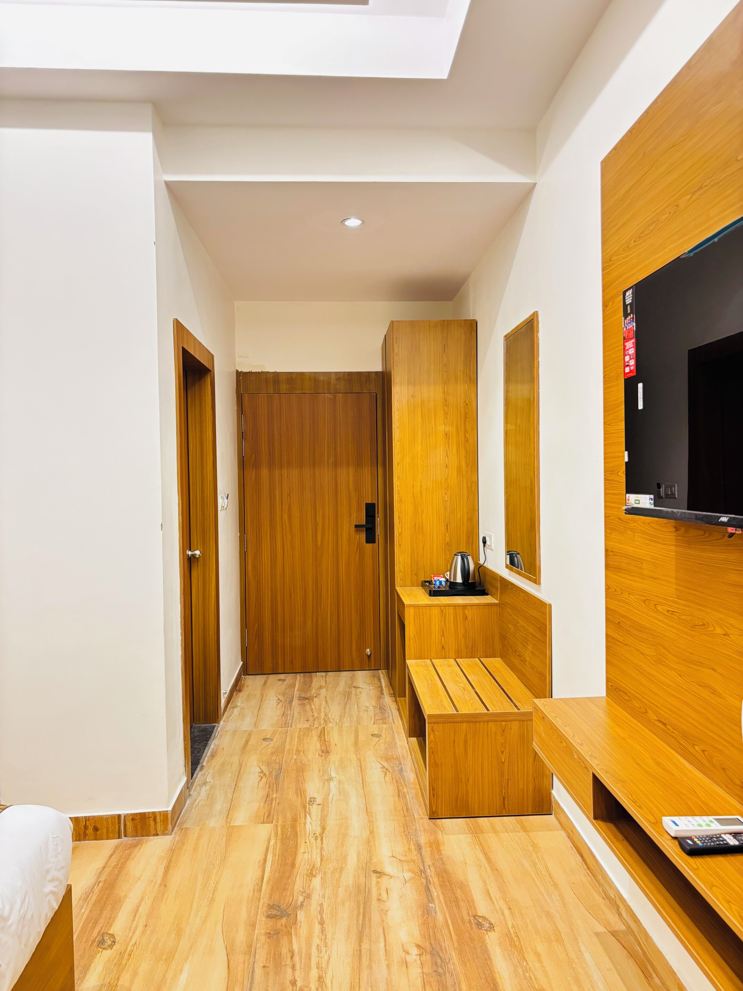 A modern hotel room with a wooden floor, a bed, a TV mounted on the wall, and a small seating area with a kettle.