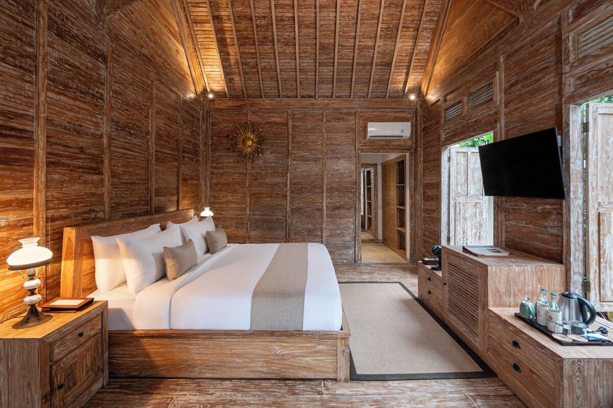 A wooden-themed bedroom with a large bed, bedside tables, a TV, and a small kitchenette area.