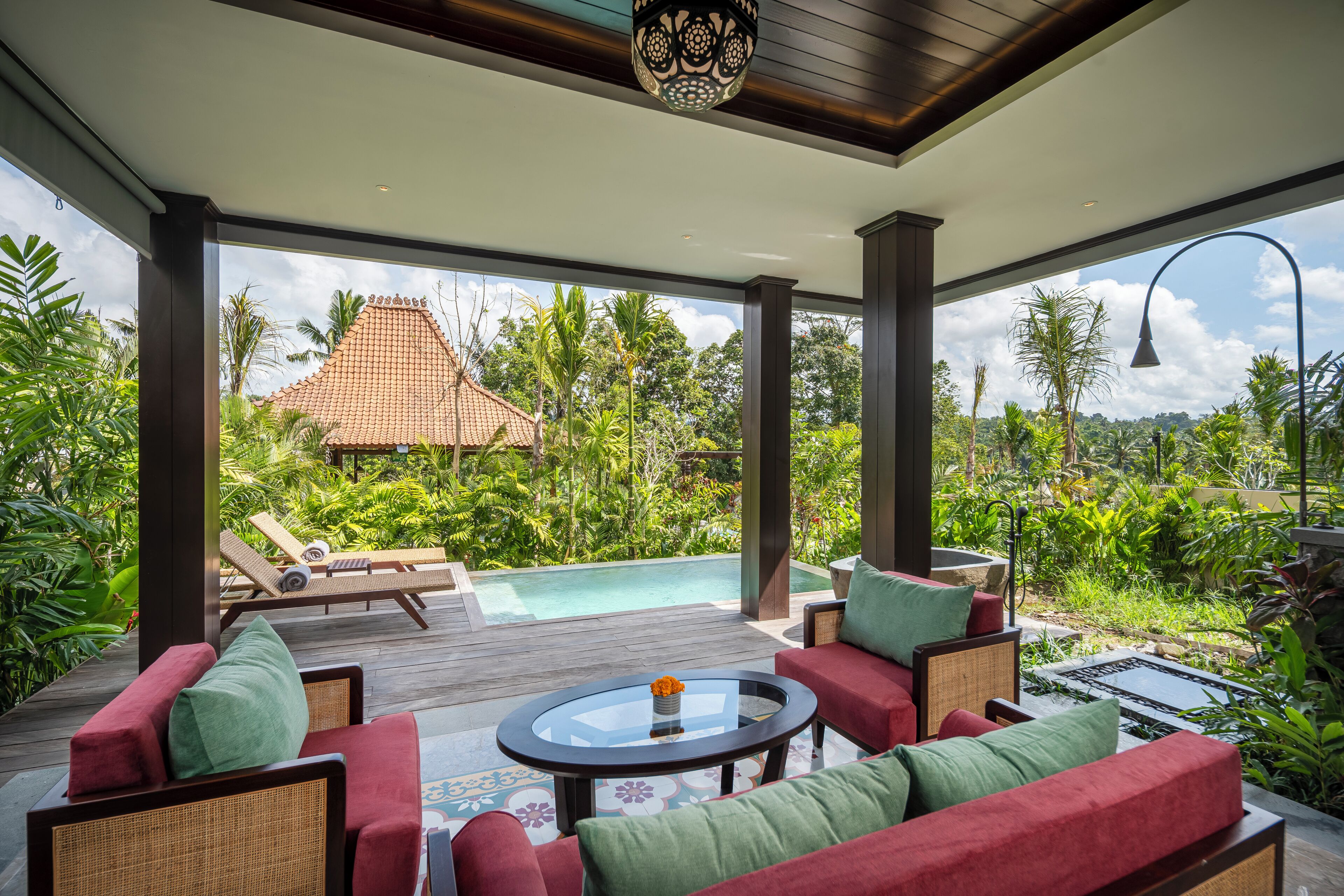 A covered outdoor area with a pool, lounge chairs, and a view of a traditional building surrounded by lush greenery.
