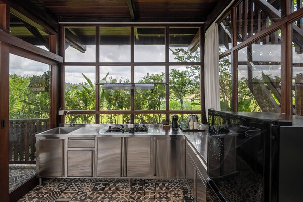 A modern kitchen with a large island, stainless steel appliances, and a view of lush greenery through large windows. - Emana Abian Taksu (Payangan)