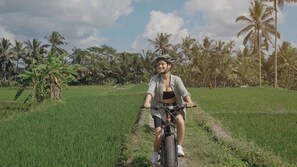 A person riding a bicycle on a path through a green field with palm trees in the background. - Emana Abian Taksu (Payangan)