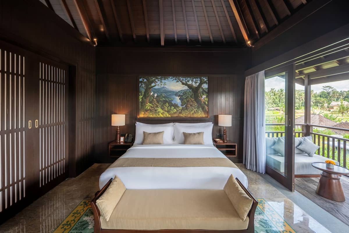 A hotel room with a large bed, a painting of a landscape, a balcony with a sofa, and a view of greenery.