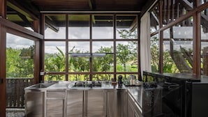 A modern kitchen with a large island, stainless steel appliances, and a view of lush greenery through large windows. - Emana Abian Taksu (Payangan)