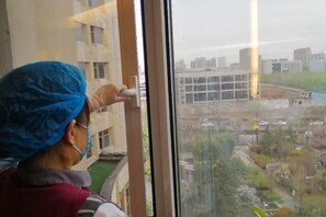 A person in a blue head covering and a red vest standing by a window, looking outside. - Yushui Hotel (Shahe)