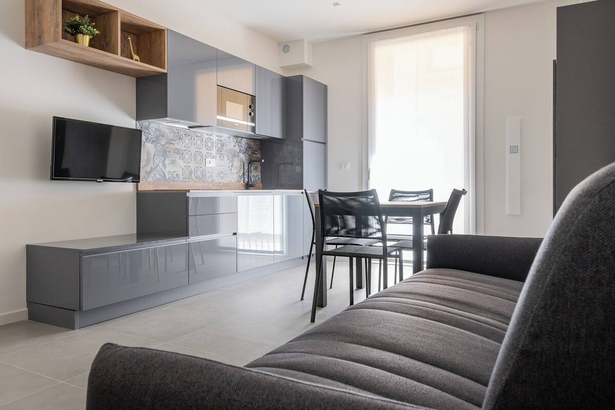 A modern living room with a sofa, a dining table with chairs, a TV mounted on the wall, and a kitchen area with cabinets and a countertop.