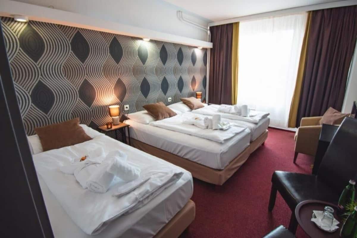 Ein Hotelzimmer mit drei Betten, einem Schreibtisch, einem Stuhl und einem großen Fenster mit Vorhängen.