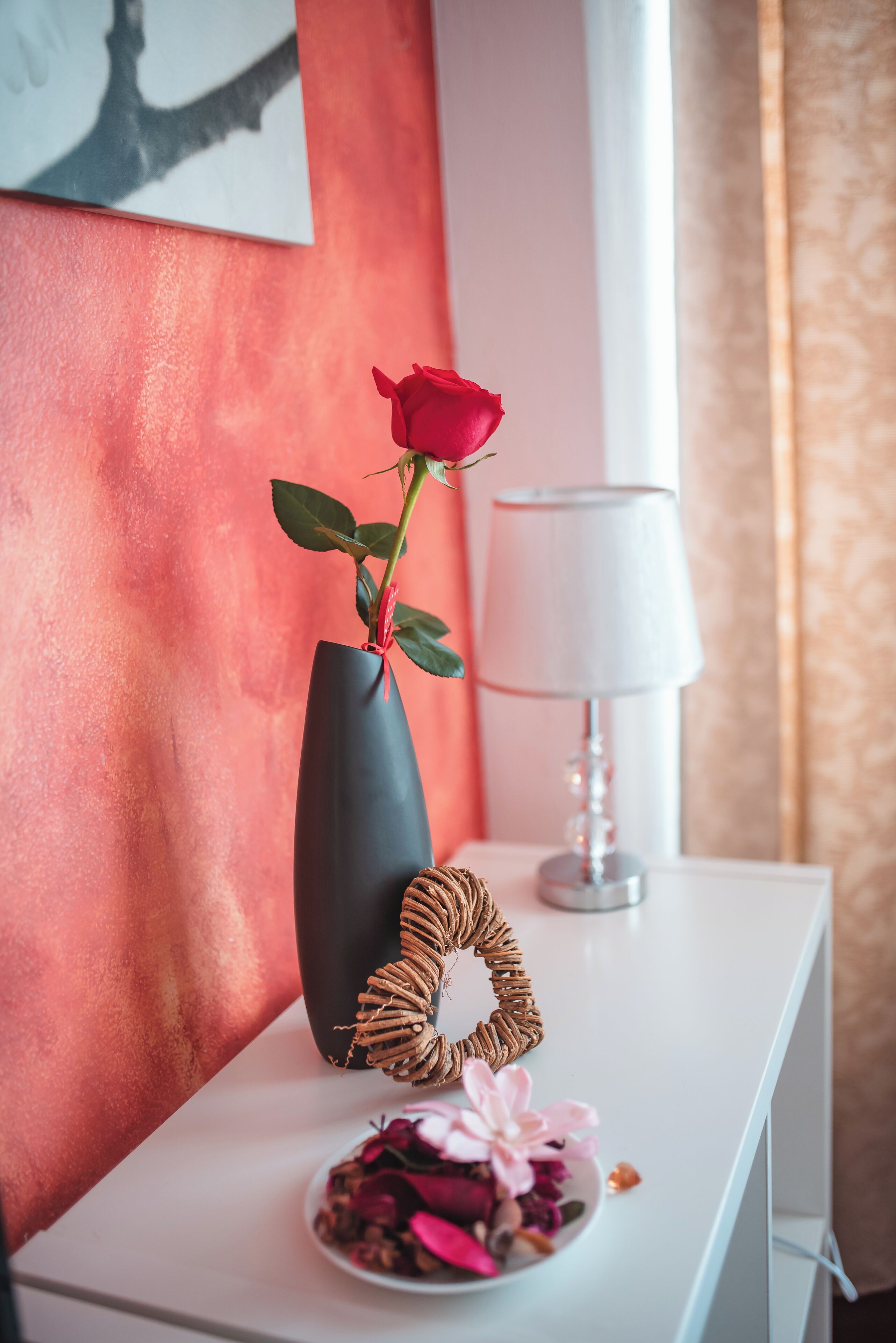 Una mesita de noche blanca con un florero oscuro que sostiene una rosa roja solitaria, un cuenco pequeño de flores secas y un adorno tejido.