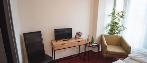 A hotel room with a bed, a desk with a television, a chair, a small table with a plant, and a window with curtains.