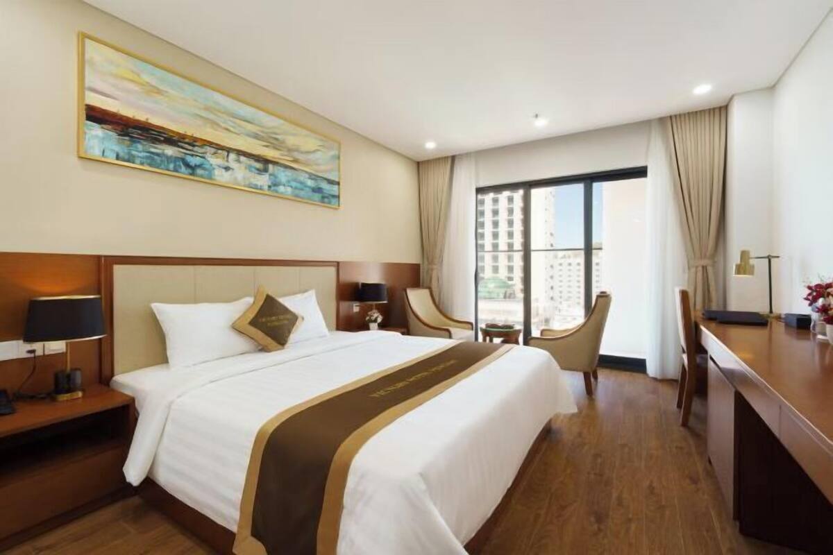 Una camera d'albergo con un letto grande, una scrivania, una sedia, una finestra con tende e un quadro appeso al muro.