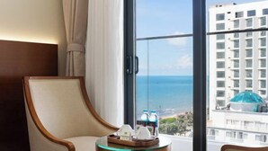 A hotel room with a large window, a chair, a small table with a tray, and a view of the ocean and buildings. - Victory Hotel Vung Tau - New Wing (Vung Tau)