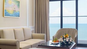 A room with a sofa, a chair, a painting, a table with a floral arrangement, and a large window with a view of the sea. - Victory Hotel Vung Tau - New Wing (Vung Tau)