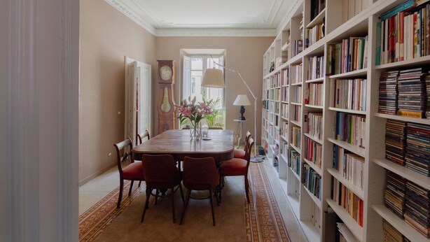 A dining room with a wooden table and chairs, a bookshelf filled with books, and a clock on the wall.