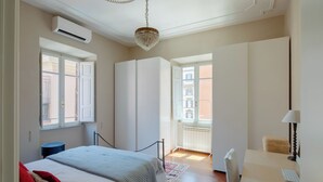 A bedroom with a bed, a desk, and a chair. There is a window, an air conditioning unit, and a chandelier.