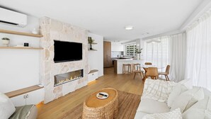 A modern living room with a stone fireplace, a flat-screen TV, and a dining area with wooden furniture. - Lookout Palm Beach - Absolute Beachfront + Views (Palm Beach)