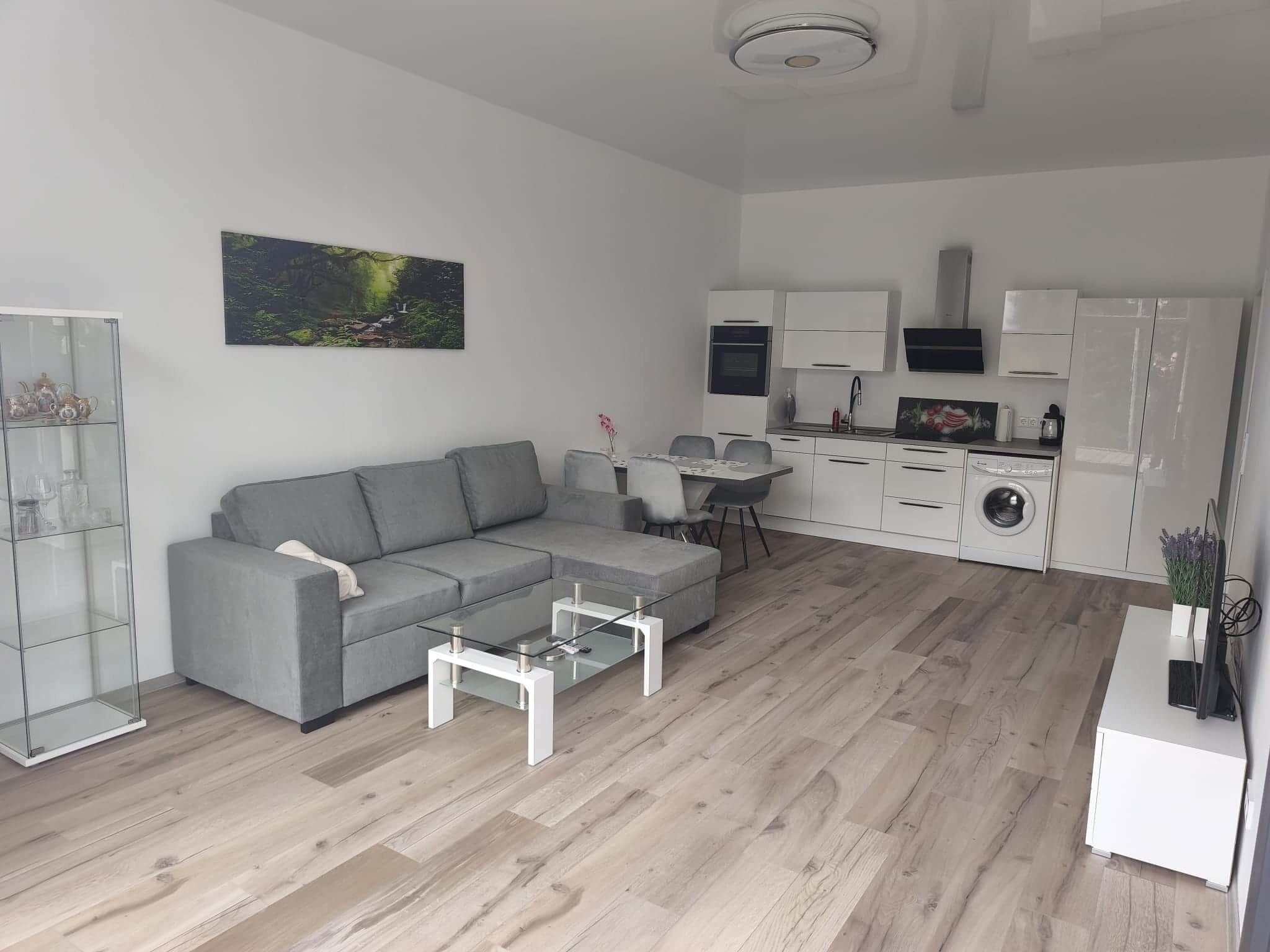 Ein modernes Wohnzimmer mit einer grauen Eckcouch, einem Glastisch und einem Essbereich mit kleinem Tisch und Stühlen. Im Hintergrund ist eine Küche mit weißen Schränken und Geräten erkennbar.