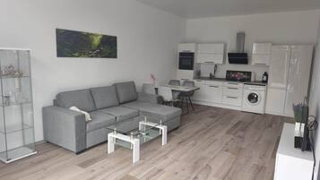 Ein modernes Wohnzimmer mit einer grauen Eckcouch, einem Glastisch und einem Essbereich mit kleinem Tisch und Stühlen. Im Hintergrund ist eine Küche mit weißen Schränken und Geräten erkennbar.