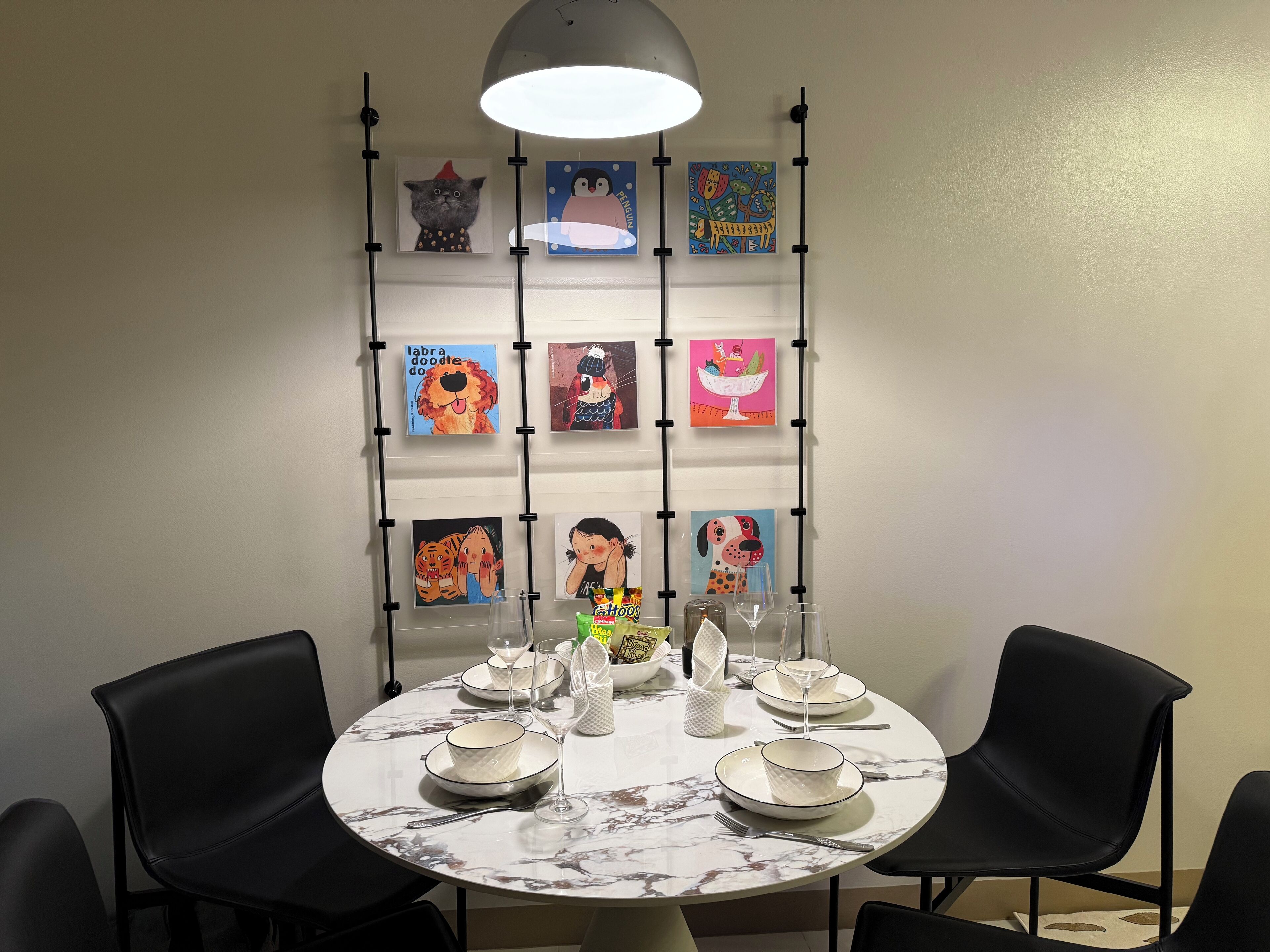 A dining area with a round table set for two, surrounded by black chairs. Above the table, there is a wall-mounted art display with various colorful paintings.