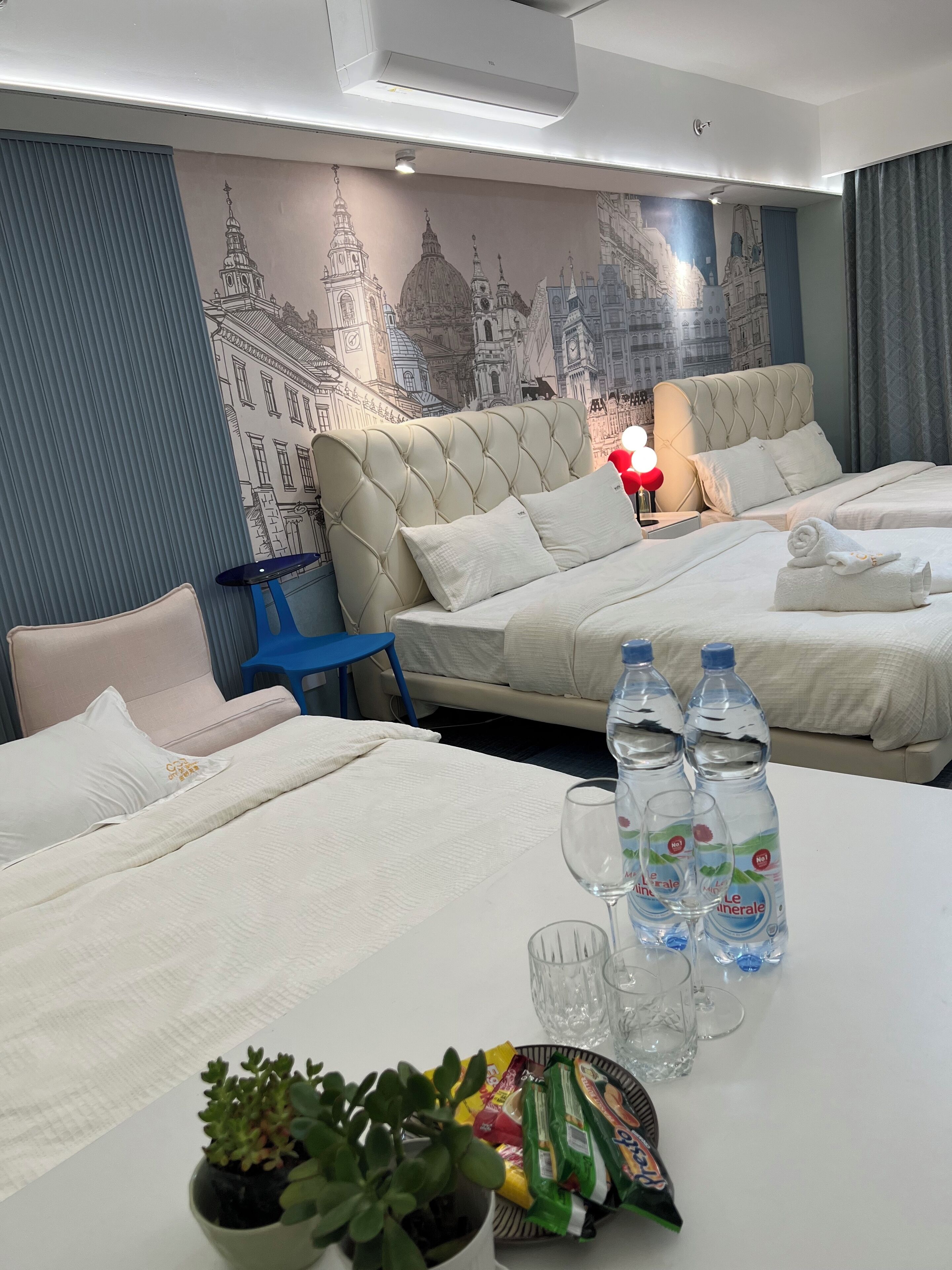 Une chambre d’hôtel avec deux lits, une table à manger avec des bouteilles d’eau et des en-cas, et une fresque représentant un paysage urbain sur le mur.