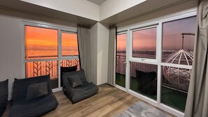 A modern living room with a large window, a balcony, and a view of a cityscape at sunset. - Luxurious European Style Modern Apartment Cross City of Dreams OKADA SOLAIRE MOA Airport (Paranaque)